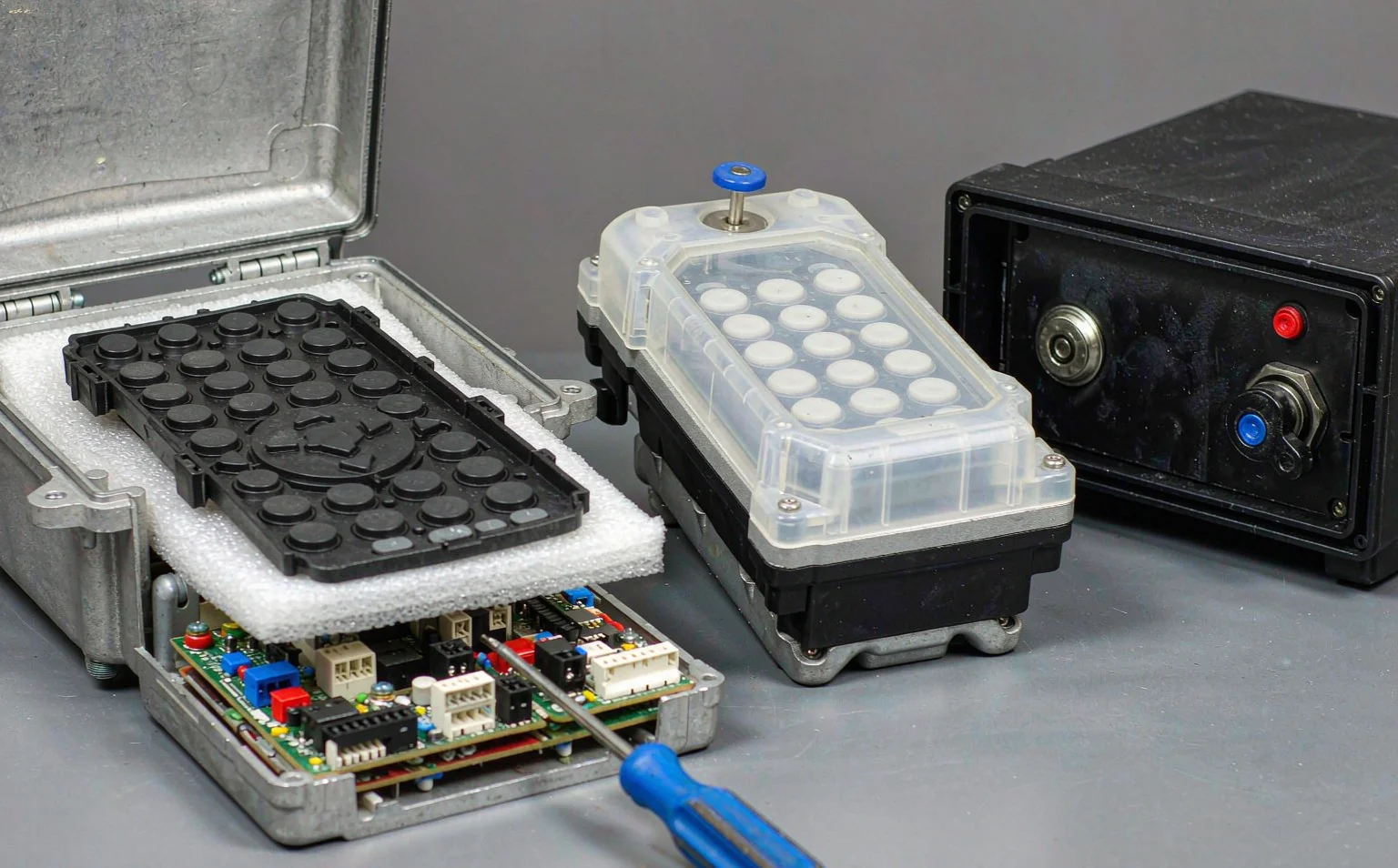 Two industrial radio remote control transmitters lying open on a bench beside a receiver with its key selector and pairing button visible