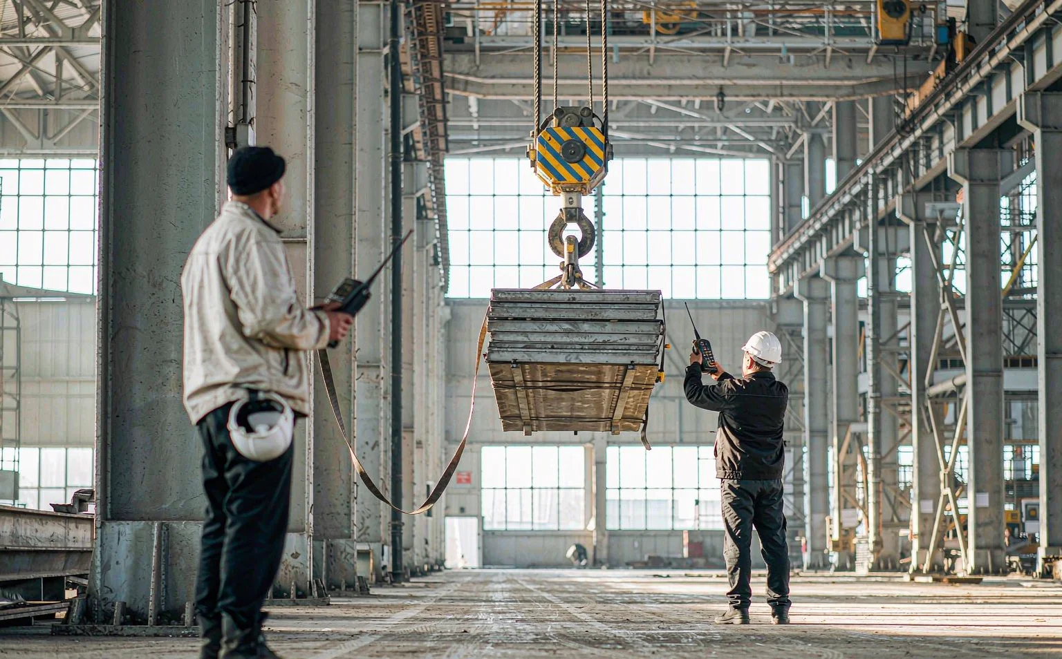 Two operators on the same crane runway each holding a radio remote control transmitter while the crane trolley moves under load