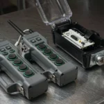 Two industrial radio remote control transmitters for one crane lying side by side on a steel workbench beside an open receiver
